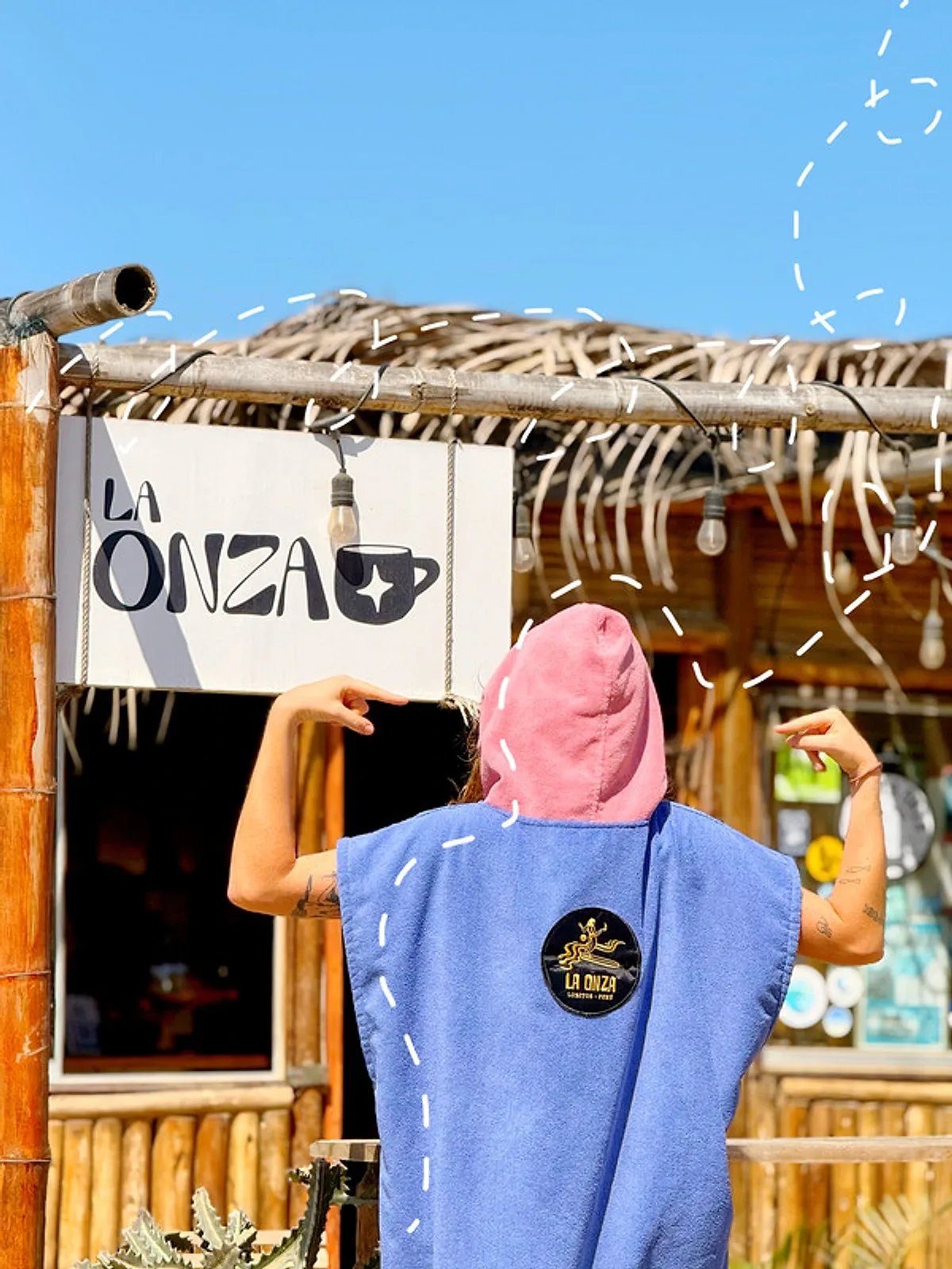 Someone wearing a blue poncho with a pink hood and the La Onza Café patch, in front of the café sign in Lobitos